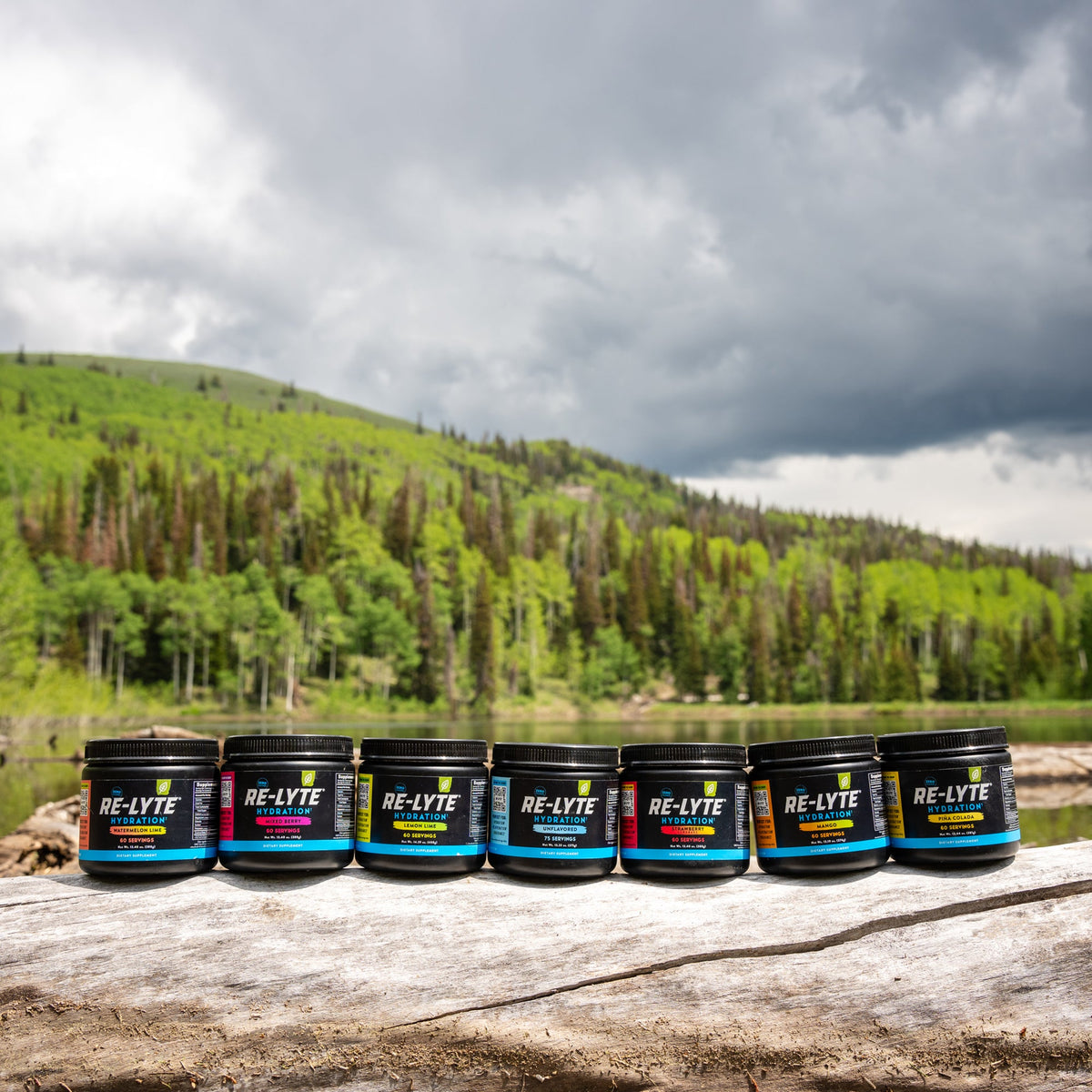 All seven Re-Lyte flavors on a log in front of a lake in the mountains of Utah. Flavors from left to right: Watermelon Lime, Mixed Berry, Lemon Lime, Unflavored, Strawberry Lemonade, Mango and Piña Colada.